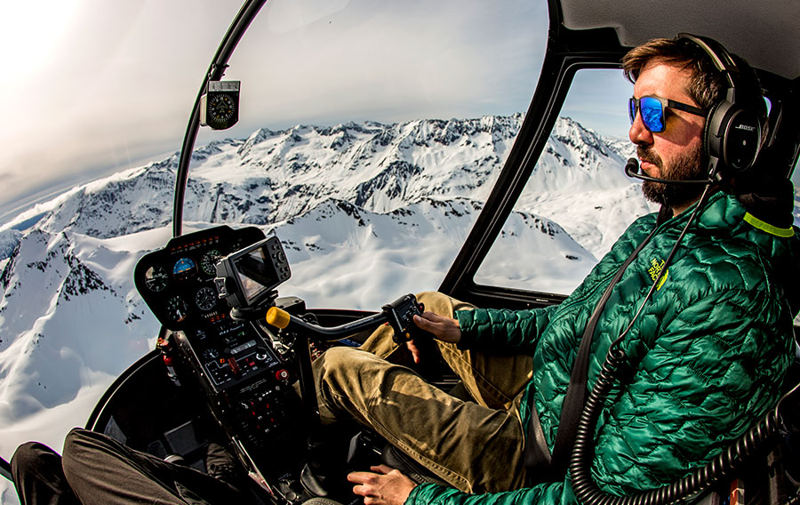 Helicopter pilot in the cockpit with snowy, Alaskan mountains behind.