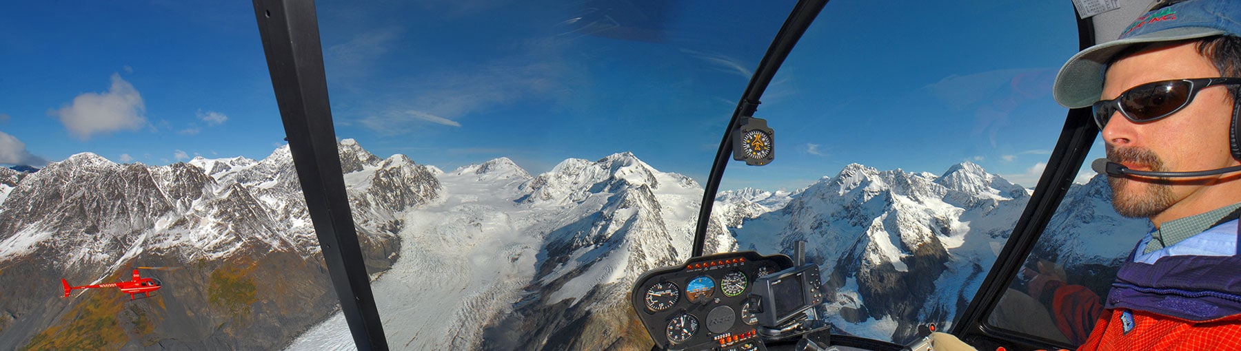 Helicopter pilot in the cockpit with snowy mountains outside.