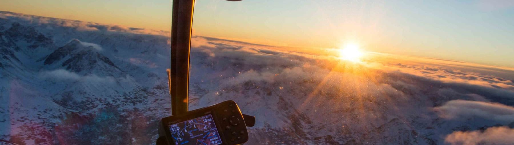 View from helicopter cockpit of sun breaking through the clouds on the horizon with snowy mountain foreground.