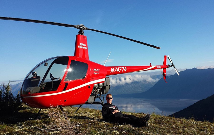 Helicopter pilot sitting next to helicopter landed on mountain ridge.