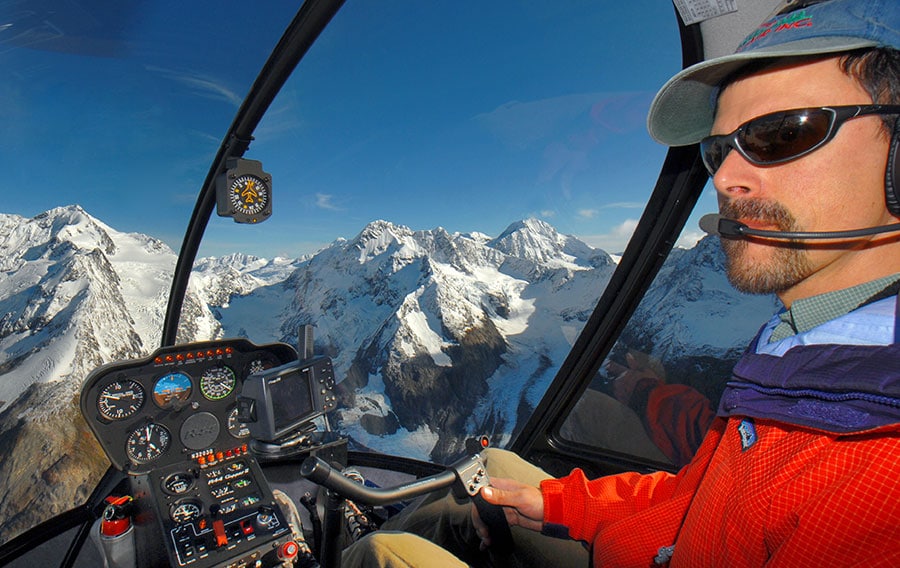 Helicopter pilot in the cockpit with snowy mountains outside.