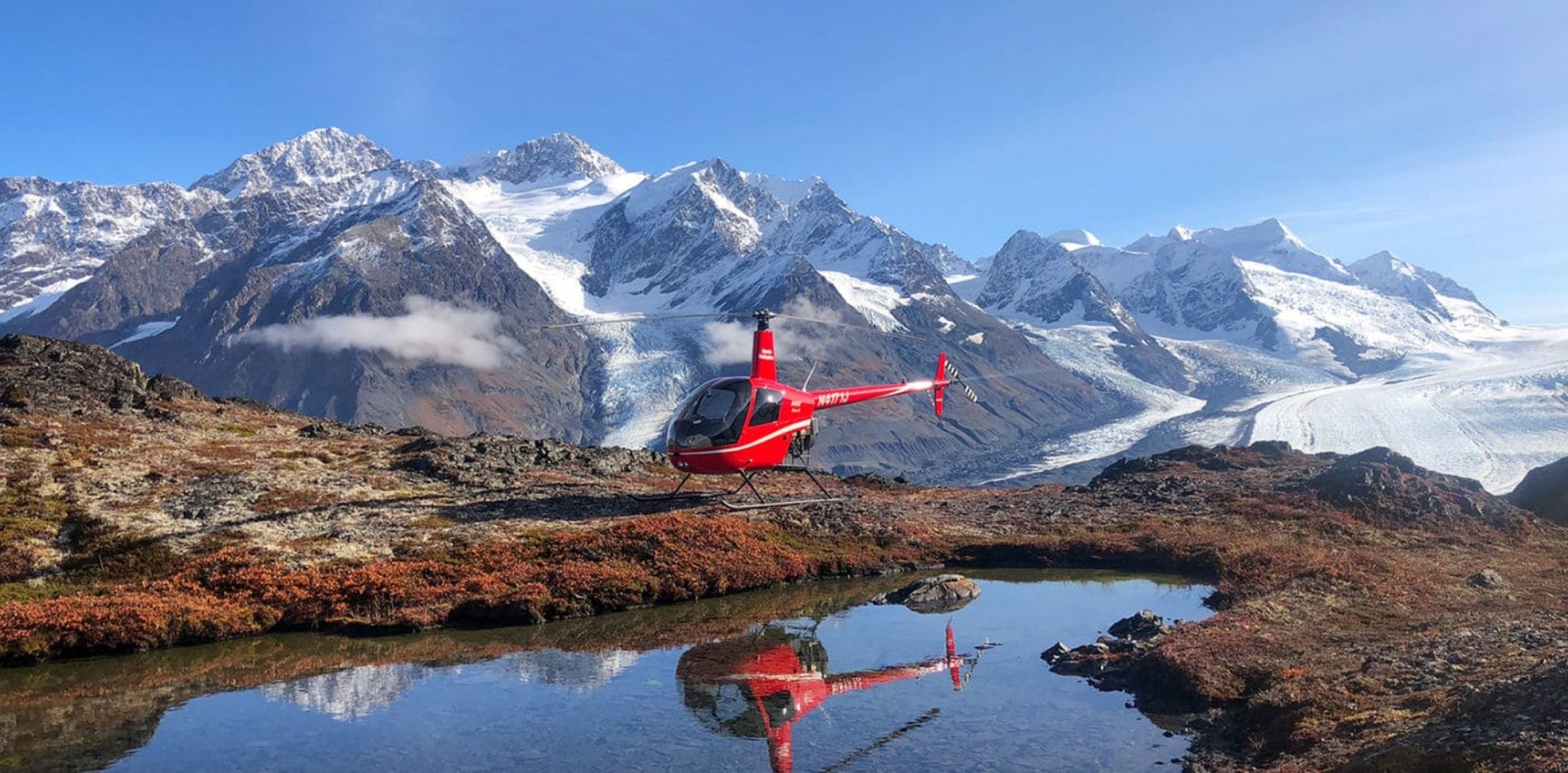 Helicopter resting on tundra next to lake with Alaskan glacier and mountains in the background.